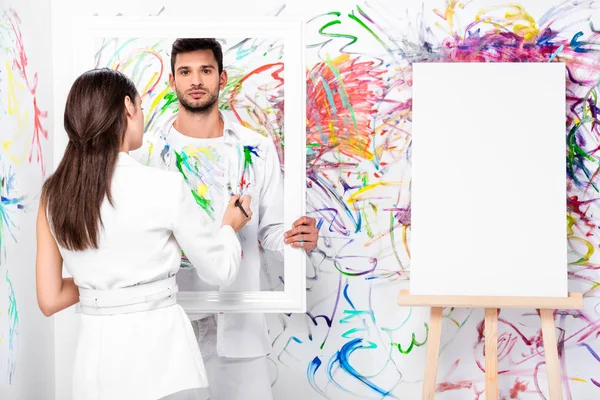 beautiful adult woman drawing on clothes while man holding frame near painted wall