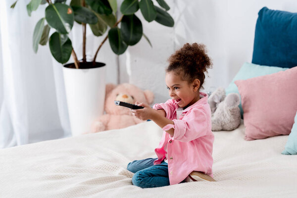 Emotional  African american child sitting on the bed with tv remote and switching the channels