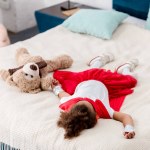 Little child in red superhero costume with teddy bear lying on bed