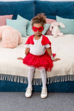 Cute little african american child in red superhero costume and mask sitting on the bed clipart