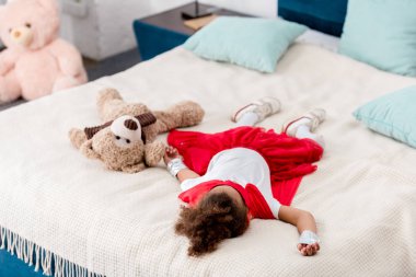 little child in red superhero costume with teddy bear lying on bed clipart