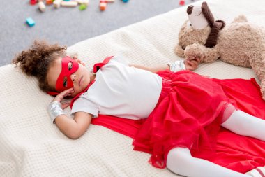 Adorable little african american child in superhero costume lying on bed with teddy bear clipart