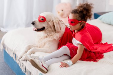 Adorable little african american kid with dog in red superhero costumes sitting on bed clipart