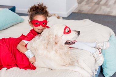 Sevimli küçük Afro-Amerikan çocuk ile köpek yatakta yatıyordu kırmızı süper kahraman kostümleri