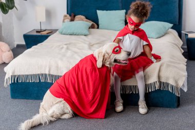 Yatak odasında süper kahraman kostümleri köpekle oynayan sevimli küçük Afro-Amerikan çocuk