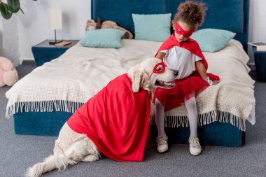 Adorable little african american kid playing with dog in superhero costumes in bedroom clipart