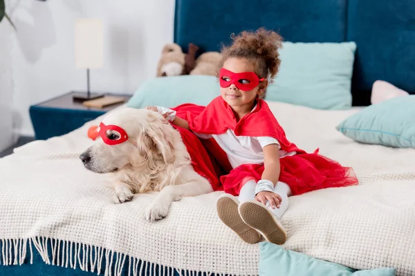 ベッドの上に座って赤いスーパー ヒーロー衣装で犬と一緒にかわいい小さなアフリカ系アメリカ人子供 — ストック写真