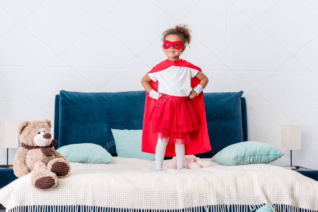 little african american kid in red superhero costume and mask with hands on hips standing on bed 