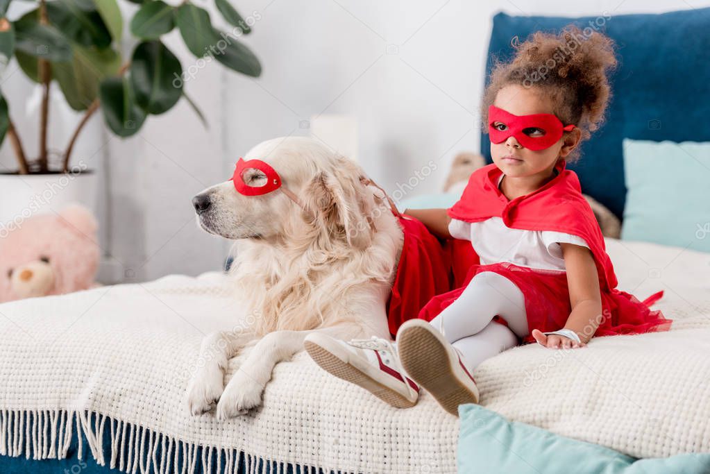 Adorable little african american kid with dog in superhero costumes sitting on bed