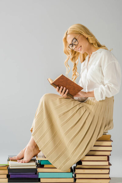 attractive smiling barefoot woman in glasses reading and sitting on books  