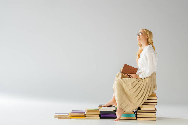 attractive dreaming barefoot woman in glasses sitting on steps made of books 