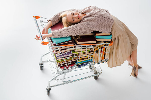 relaxed woman laying on pile of vintage books with multicolored covers in shopping cart isolated on grey