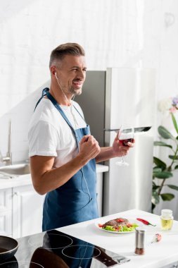 smiling man with glass of wine listening to music at kitchen clipart