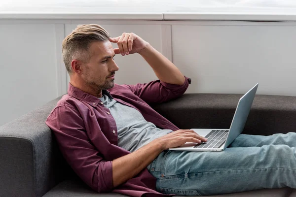 Вдумливий Чоловік Лежить Дивані Працює Ноутбуці Домашньому Офісі — Безкоштовне стокове фото