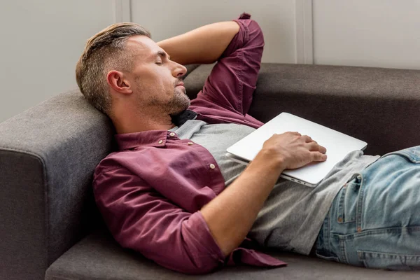 男子躺在沙发上与笔记本电脑和睡在家里的办公室 — 图库照片