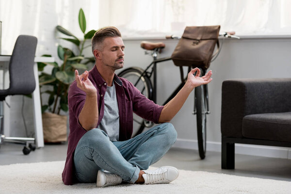 концентрированный мужчина, сидящий на полу и практикующий йогу в домашнем офисе
 