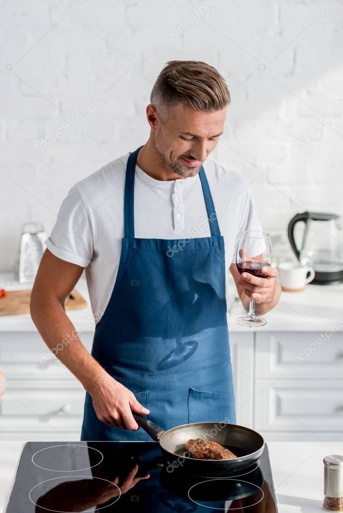  smiling man in apron with glass of wine and roasting steak on pan