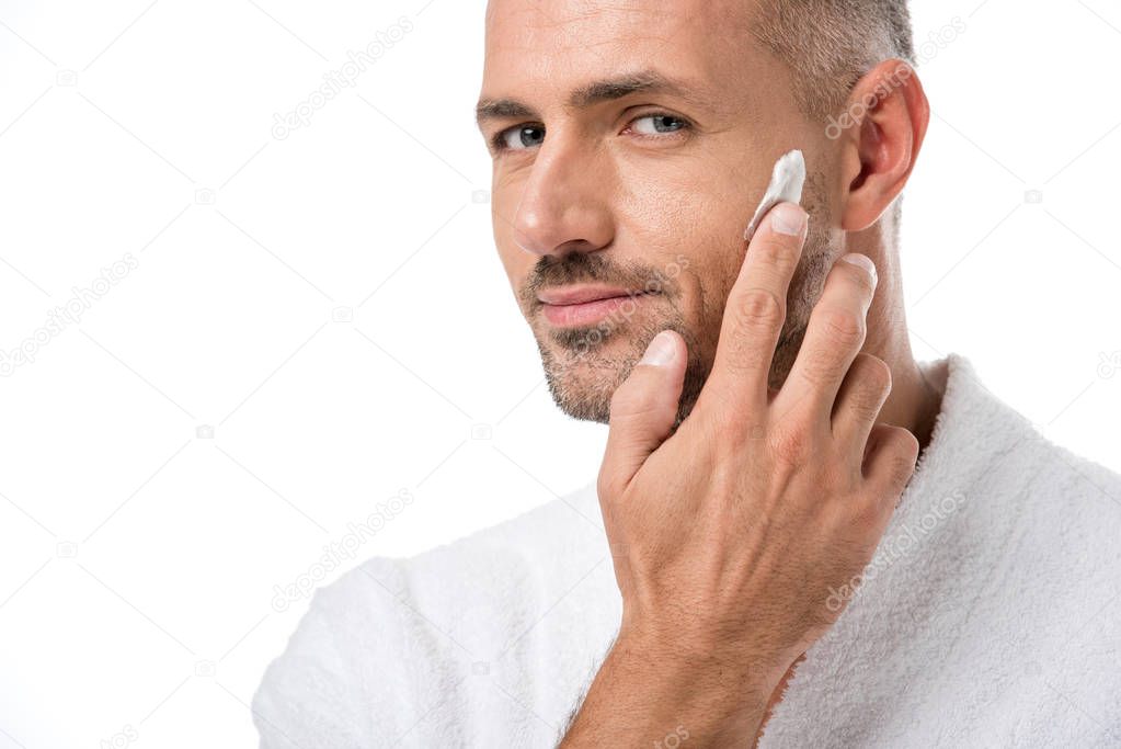 adult handsome man in bathrobe applying beauty cream on face isolated on white