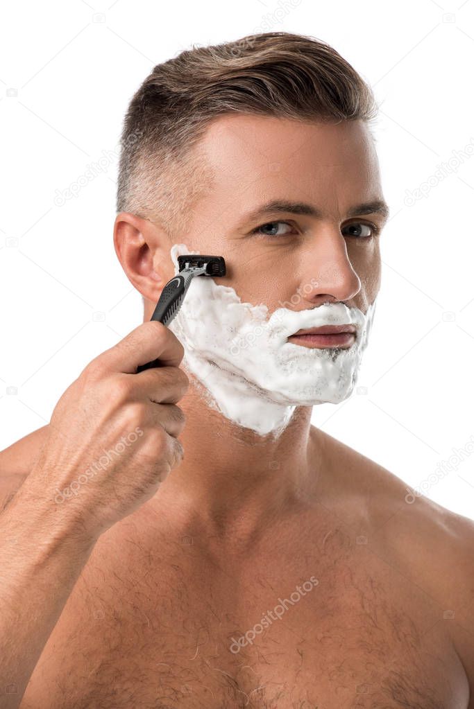 focused adult man with foam on face shaving with razor isolated on white