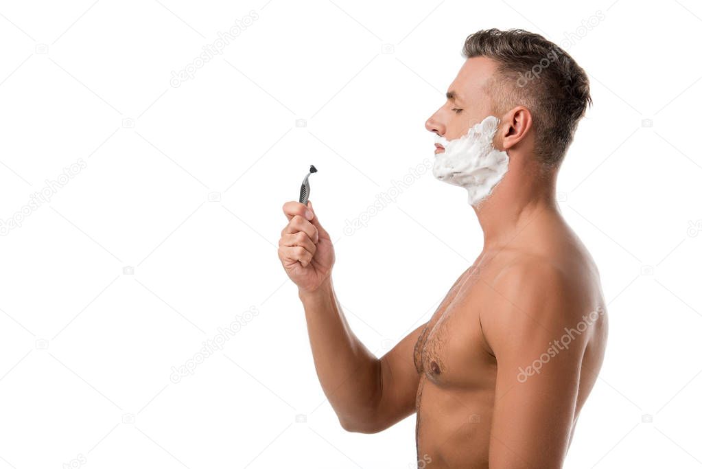 side view of adult man with shaving foam on face holding razor isolated on white