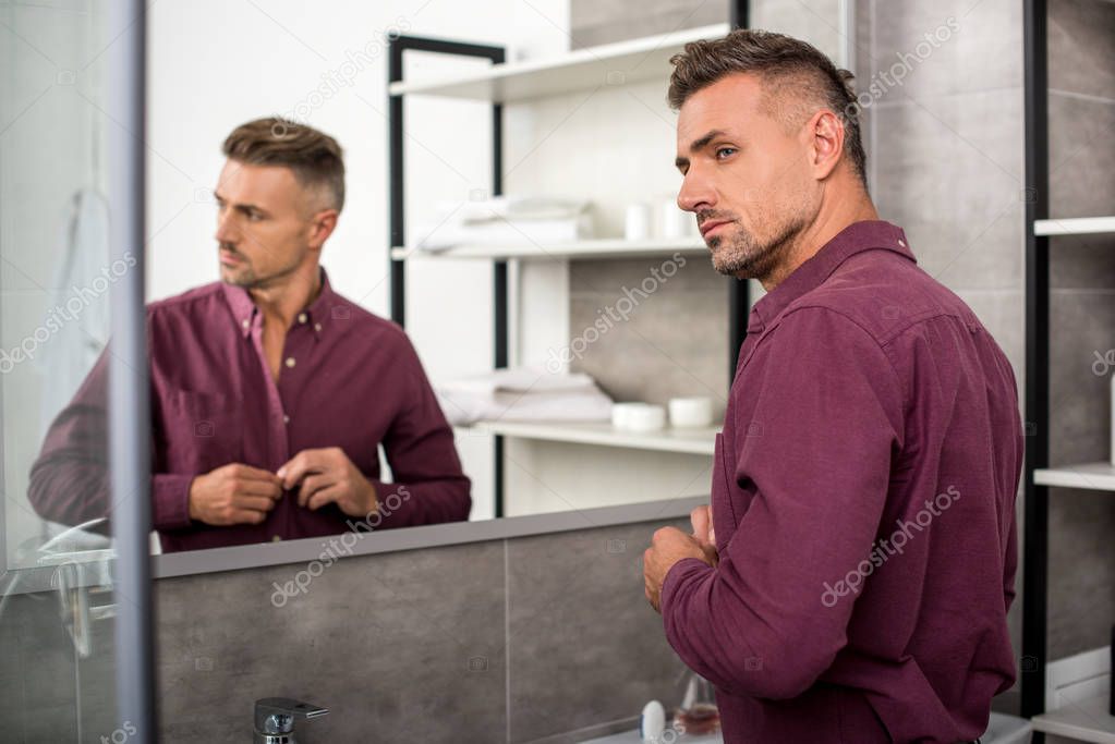 selective focus of adult businessman buttoning up shirt in bathroom at home