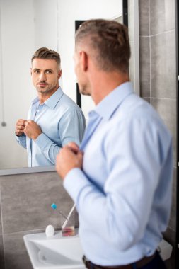 rear view of adult businessman looking at mirror while buttoning up blue shirt in bathroom at home clipart