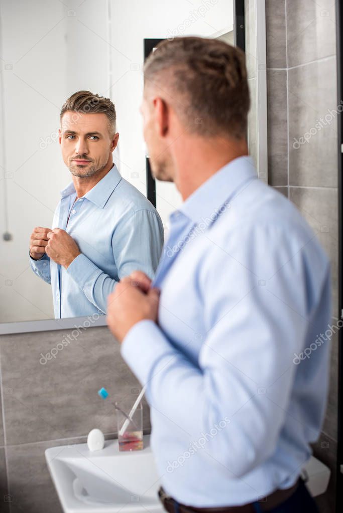 rear view of adult businessman looking at mirror while buttoning up blue shirt in bathroom at home