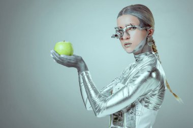 yeşil elma tutarak ve izole gri, geleceğin teknoloji kavramı üzerinde kamera bakarak cyborg yan görünüm