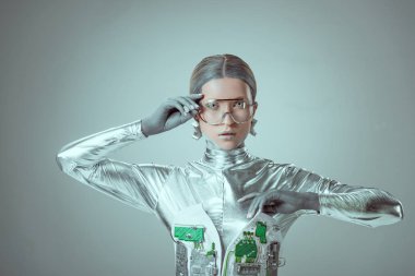 young woman robot in futuristic eyeglasses looking at camera isolated on grey, future technology concept clipart