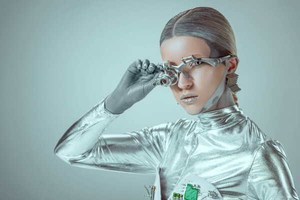 futuristic silver cyborg adjusting eye prosthesis and looking at camera isolated on grey, future technology concept  