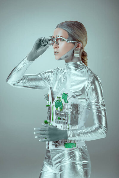 futuristic silver cyborg adjusting eye prosthesis and looking away isolated on grey, future technology concept 