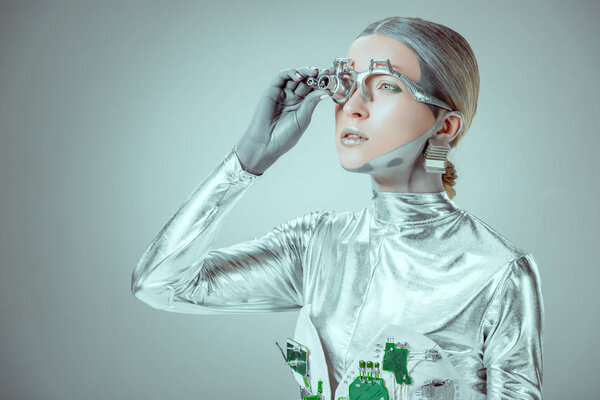 futuristic silver robot adjusting eye prosthesis and looking away isolated on grey, future technology concept  