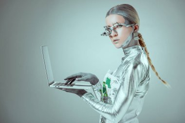 Gümüş robot dizüstü holding ve izole gri, geleceğin teknoloji kavramı üzerinde kamera bakıyor yan görünüm
