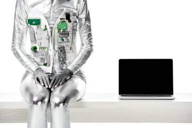 cropped image of robot sitting on table near laptop with blank screen isolated on white, future technology concept clipart