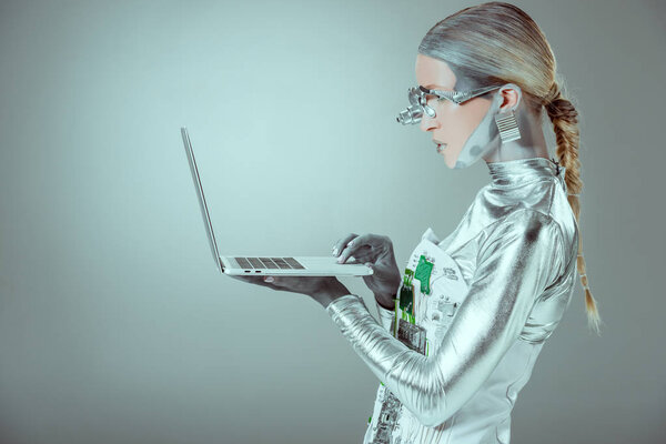 side view of silver robot holding laptop isolated on grey, future technology concept