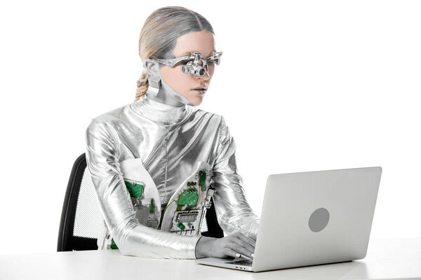 silver robot sitting at table and working with laptop isolated on white, future technology concept