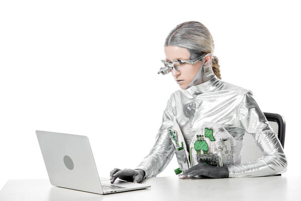 silver robot sitting at table and using laptop isolated on white, future technology concept