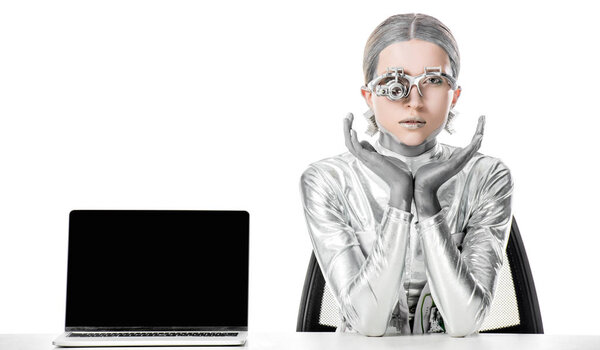 Silver robot sitting at table near laptop with blank screen isolated on white, future technology concept
