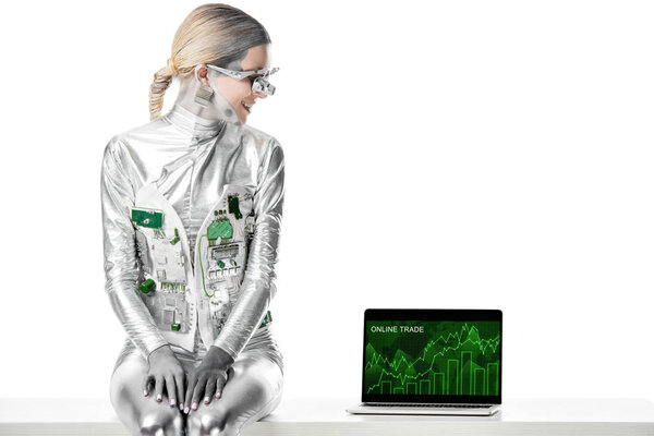 Улыбающийся серебряный робот сидит на столе и смотрит на ноутбук с онлайновым торговым устройством, изолированным на белой, будущей концепции технологии
