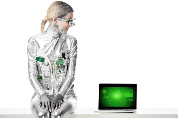 smiling silver robot sitting on table and looking at laptop with business appliance isolated on white, future technology concept