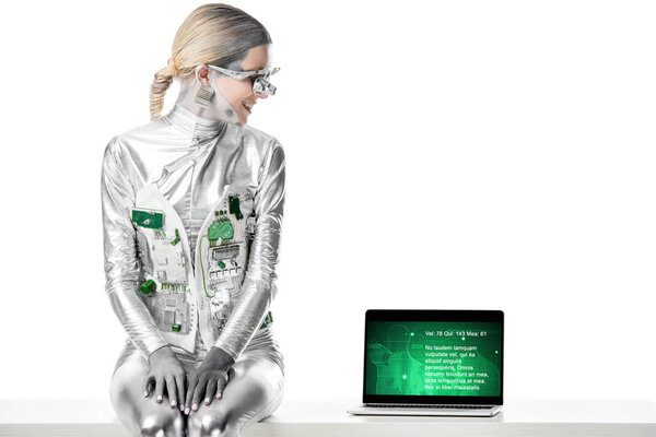 улыбающийся серебряный робот сидит на столе и смотрит на ноутбук с медицинским устройством, изолированным на белой, будущей концепции технологии
