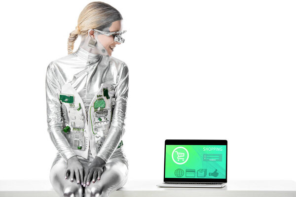 Улыбающийся серебряный робот сидит на столе и смотрит на ноутбук с устройством для покупок, изолированным на белой концепции технологии будущего
