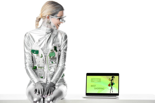 smiling silver robot sitting on table and looking at laptop with best shopping appliance isolated on white, future technology concept