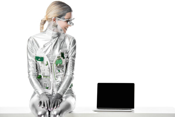 smiling silver robot sitting on table and looking at laptop with blank screen isolated on white, future technology concept