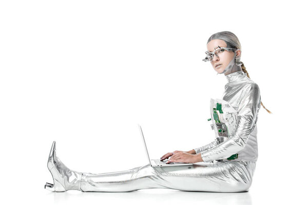 side view of silver robot sitting with laptop and looking at camera isolated on white, future technology concept