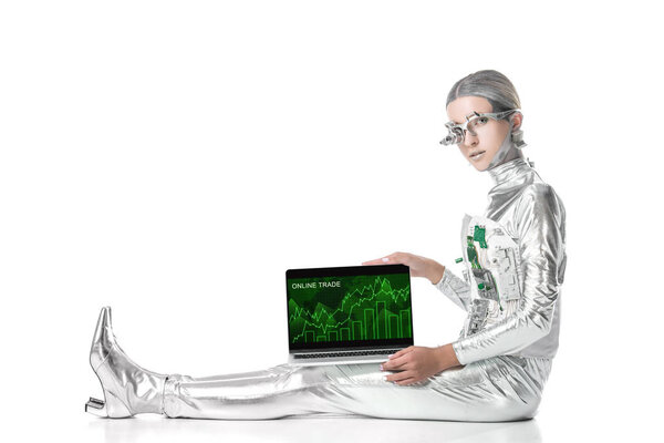 silver robot sitting and showing laptop with online trade appliance isolated on white, future technology concept