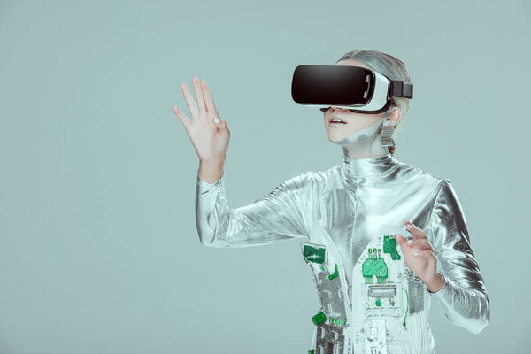 удивленный серебряный робот, трогающий что-то с помощью гарнитуры виртуальной реальности, изолированной на серой концепции технологии будущего
