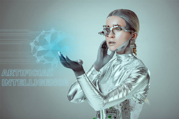 futuristic silver cyborg touching digital data with "artificial intelligence" lettering isolated on grey, future technology concept  
