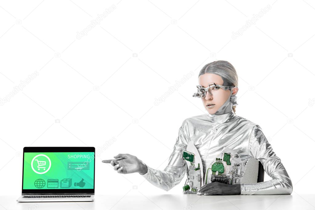 silver robot sitting at table and pointing on laptop with shopping appliance isolated on white, future technology concept