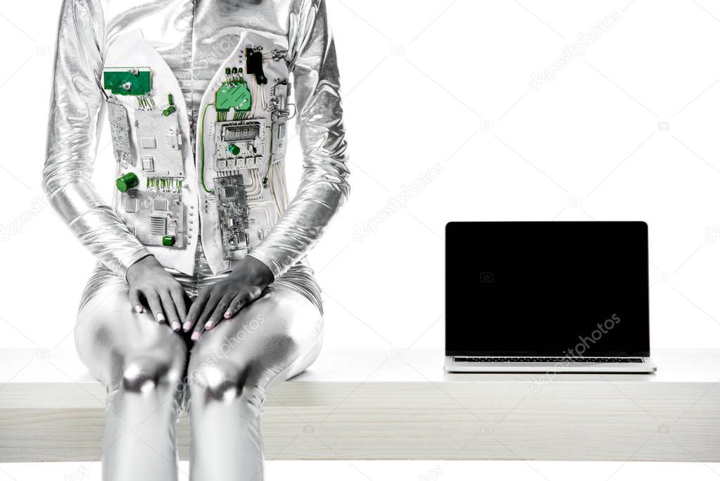 cropped image of robot sitting on table near laptop with blank screen isolated on white, future technology concept
